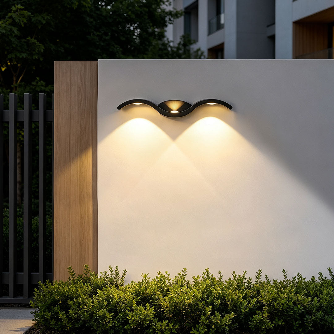 Wave wall sconce installed on a modern outdoor wall above greenery with warm dual-beam illumination.