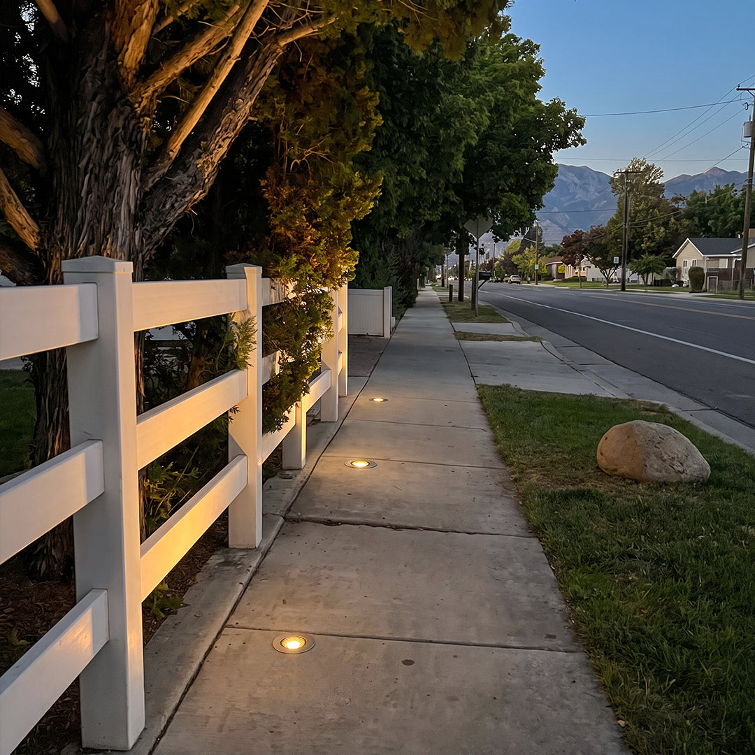 Well lights in concrete line a residential sidewalk next to a white fence, providing subtle pathway lighting at dusk.