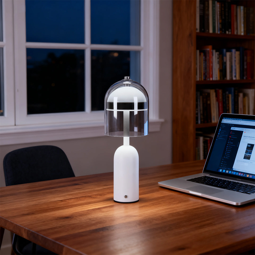 A white glass shade table lamp used as ambient light on a wooden desk with a laptop next to a window at dusk. 