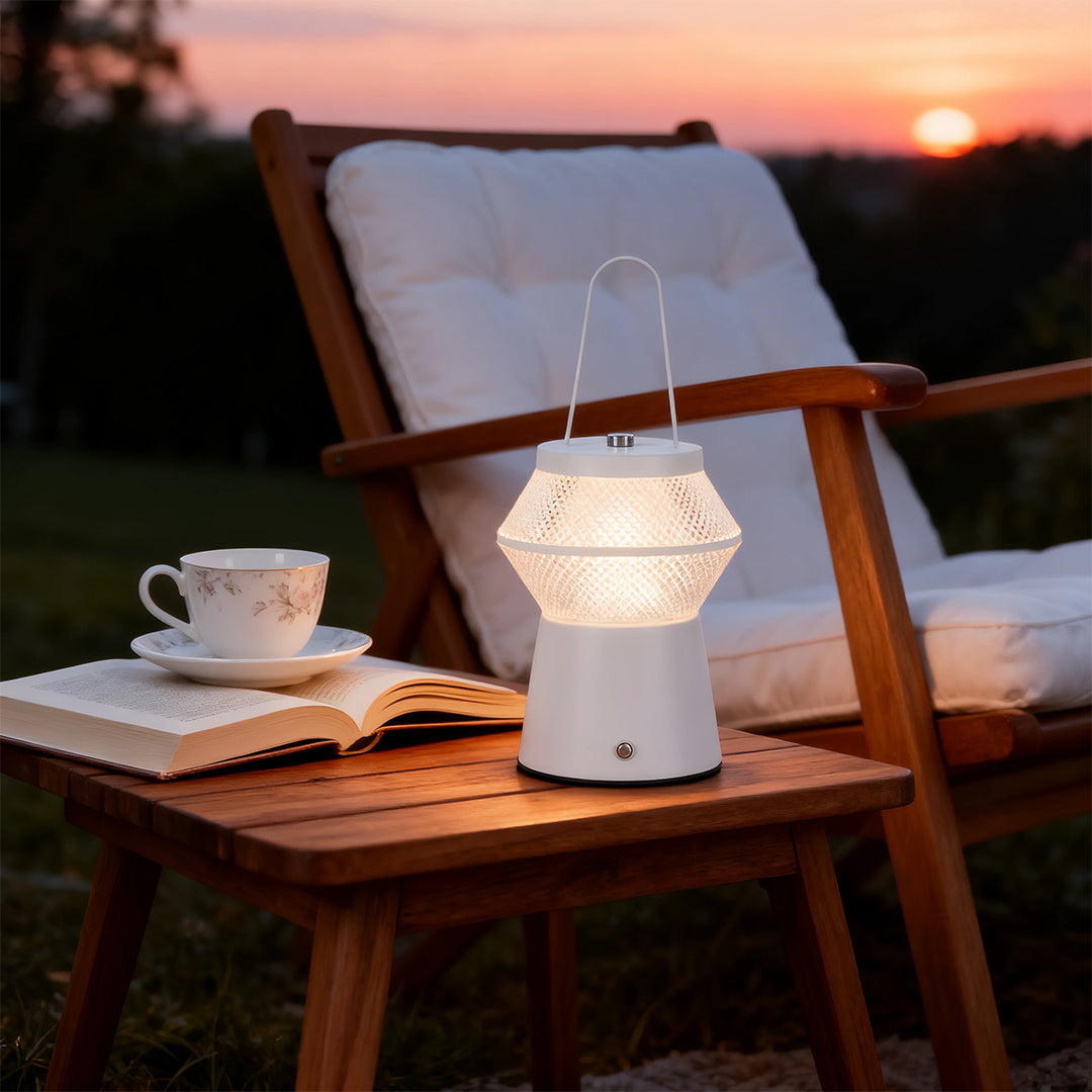 White led outdoor solar lantern with a textured shade placed on a wooden side table next to a relaxing chair at sunset. 
