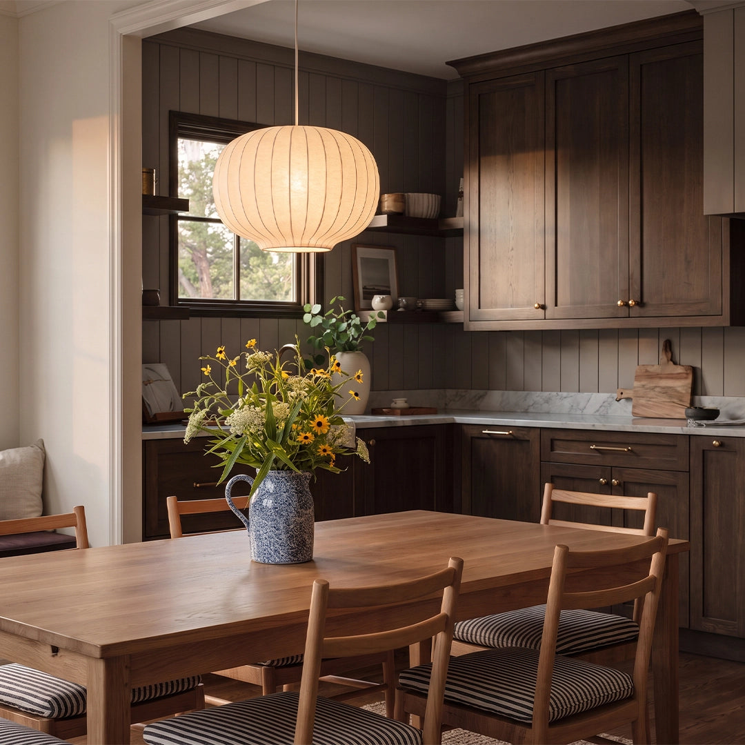 White modern chandelier suspended above a wooden dining table in a warm kitchen dining space with a pleated round shade.