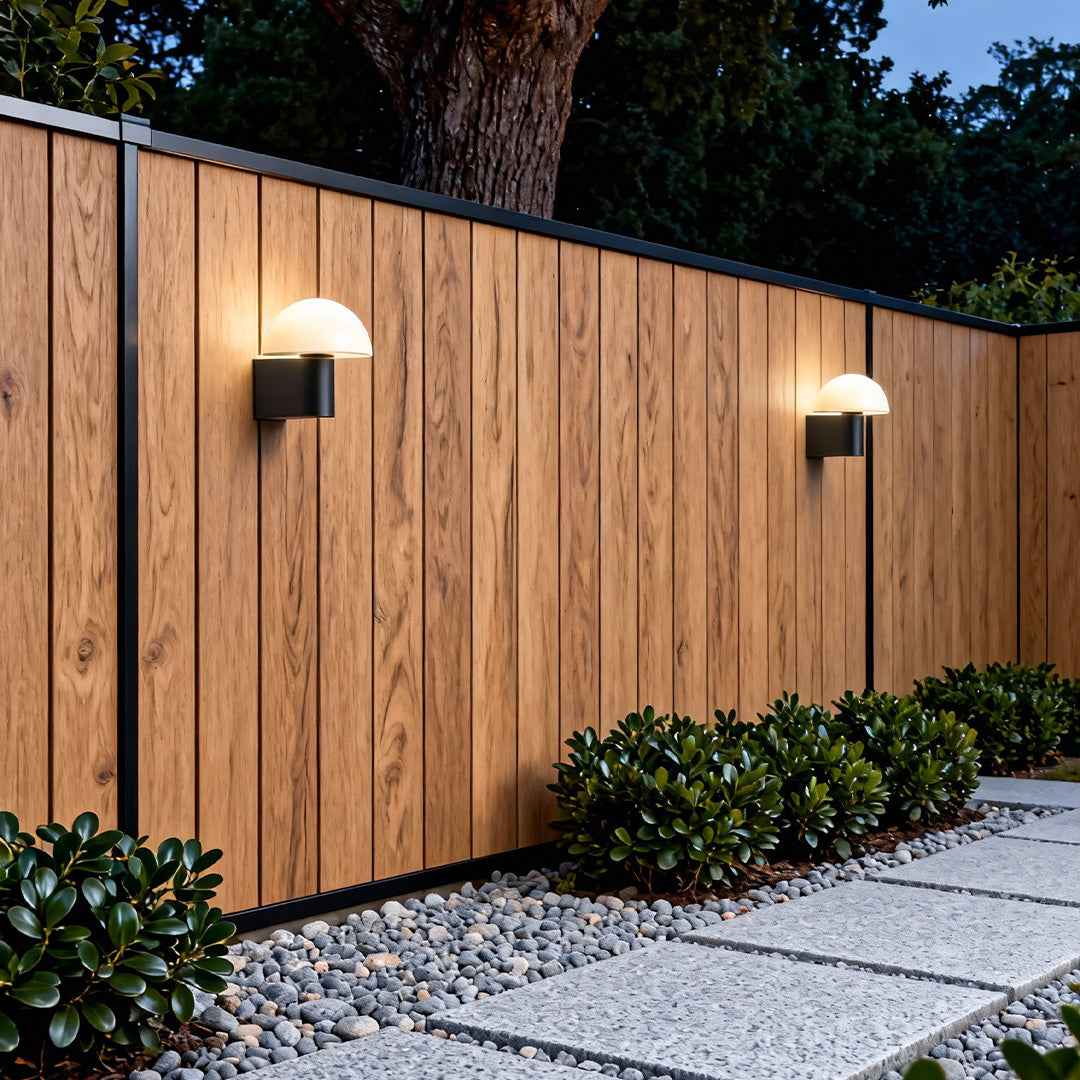 Outdoor wall light sconce installed along a wooden fence, providing warm pathway illumination in a modern garden setting.