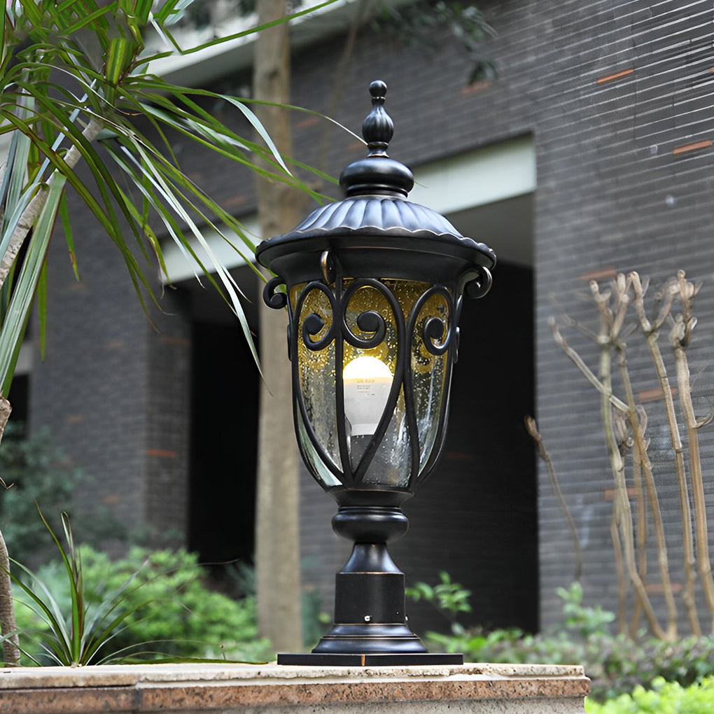 Black outdoor pillar light with clear glass panels, showcasing a warm bulb glow in a garden.