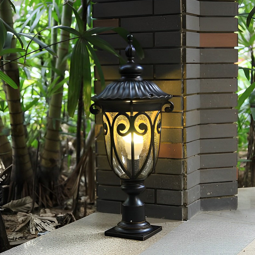 Black outdoor pillar light with decorative design, placed beside lush greenery and brick wall.