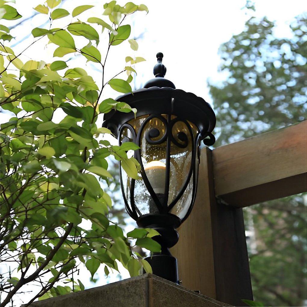Close-up of a black outdoor pillar light with ornate details, surrounded by green foliage.