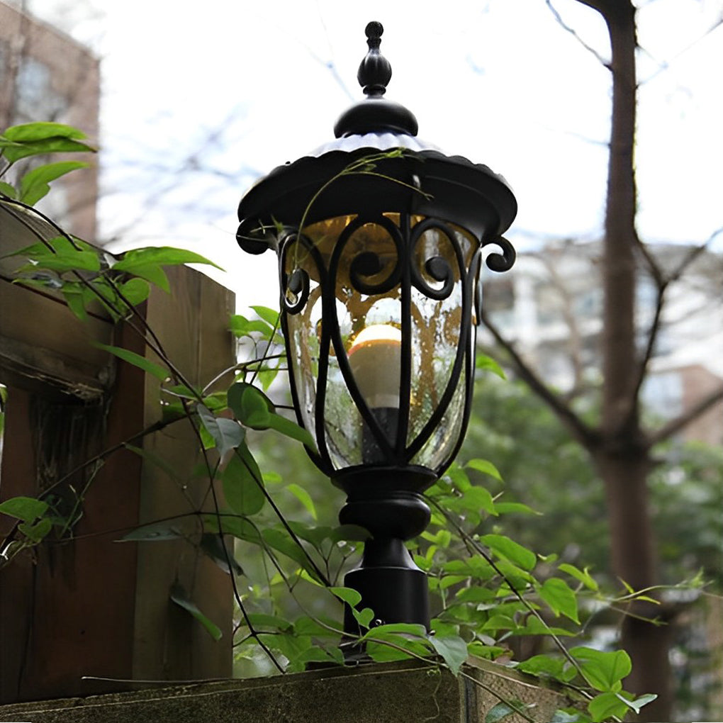 Black outdoor pillar light with vintage design, installed in a garden with plants and trees.