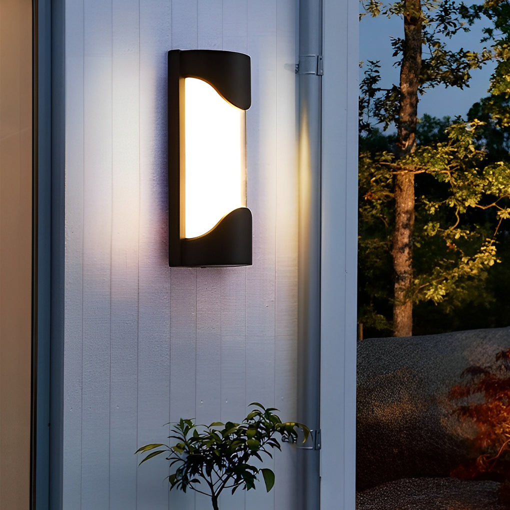 Black outdoor wall light with curved design on white wall near green plants.
