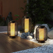 Lantern metal outdoor light with cylindrical design, placed on pebbles beside garden plants for warm ambient glow.