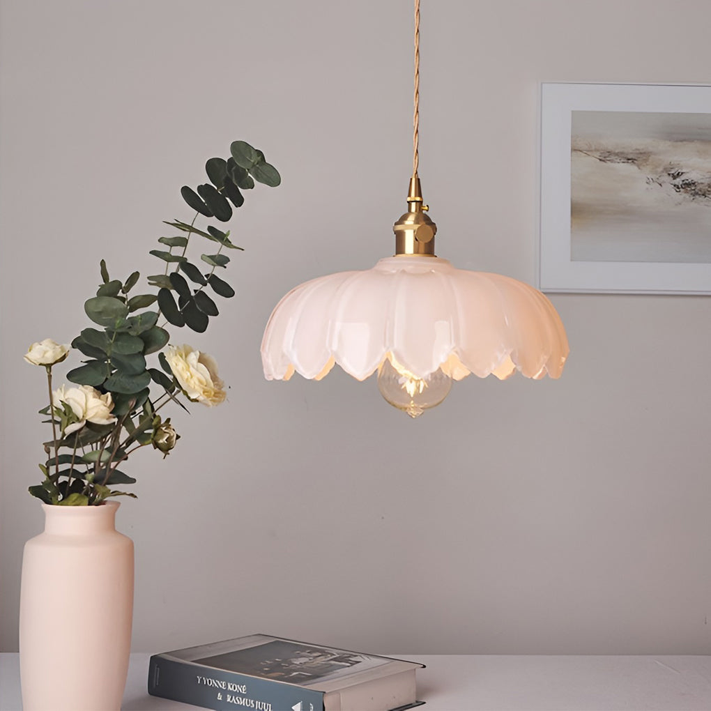 Glass pendant light with soft pink scalloped shade and brass fixture, glowing warmly in a minimalist indoor setting.