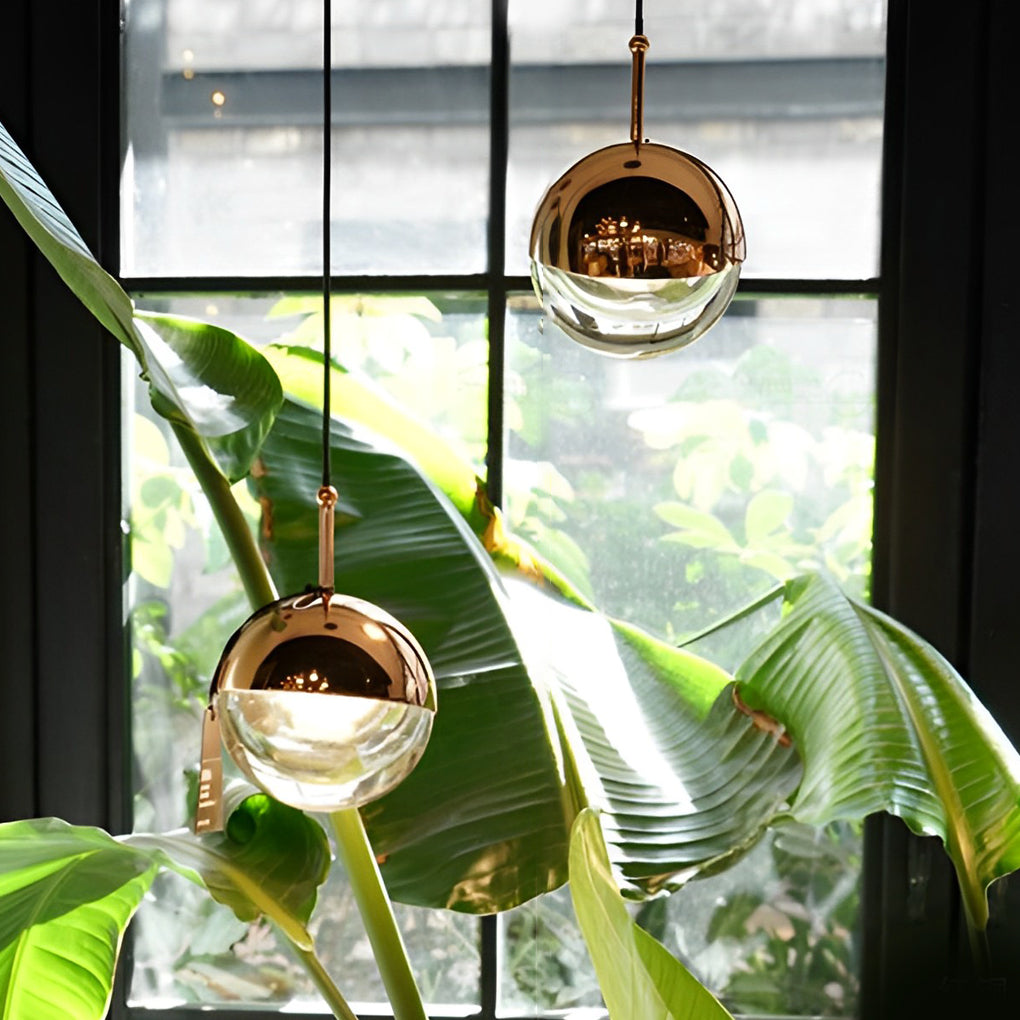 Gold globe pendant light hanging in front of a window with large green tropical plant leaves.