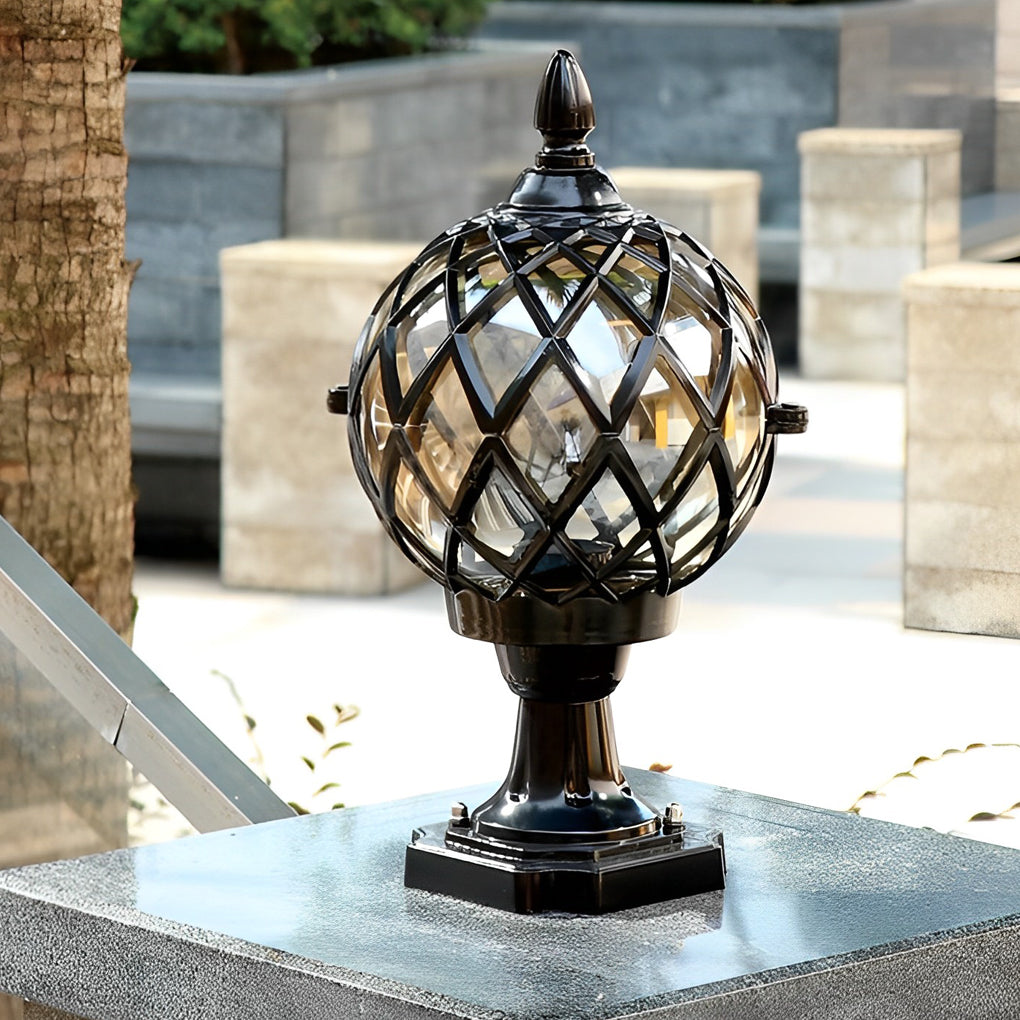 Modern home pillar light in black and gold, standing on a concrete base near a tropical garden setting.