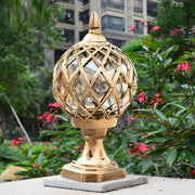 Outdoor home pillar light with a decorative gold globe, illuminating a lush garden area with vibrant plants.
