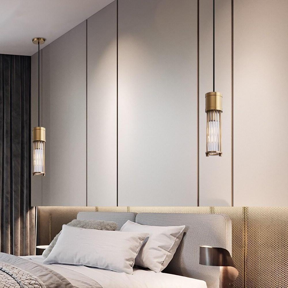 Minimal gold cylinder pendant light hanging over upholstered bed.