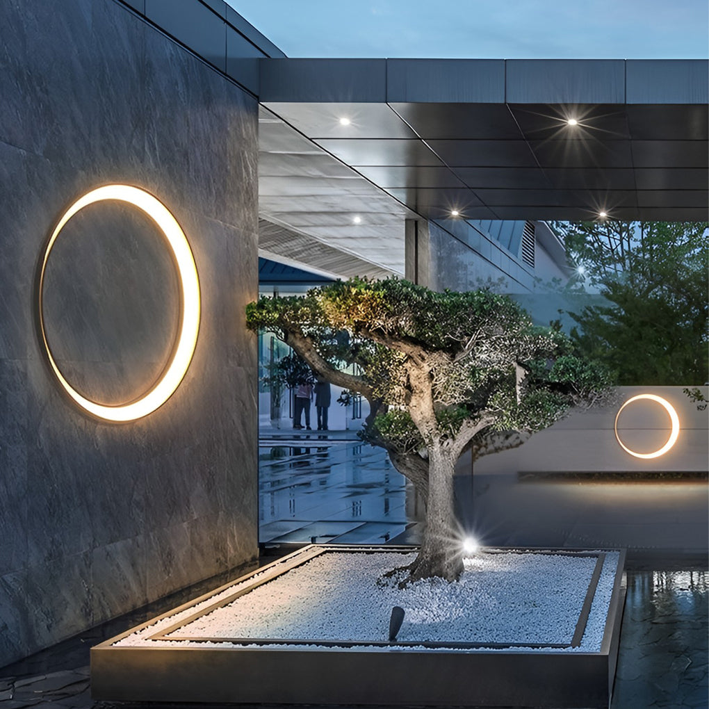 Outdoor cylinder wall light accenting a courtyard wall with trees and reflective surfaces, adding modern architectural ambience at night.