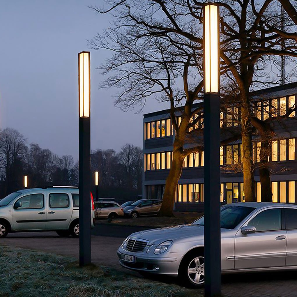 Row of black outdoor bollard lights illuminating an office parking lot near parked silver cars.