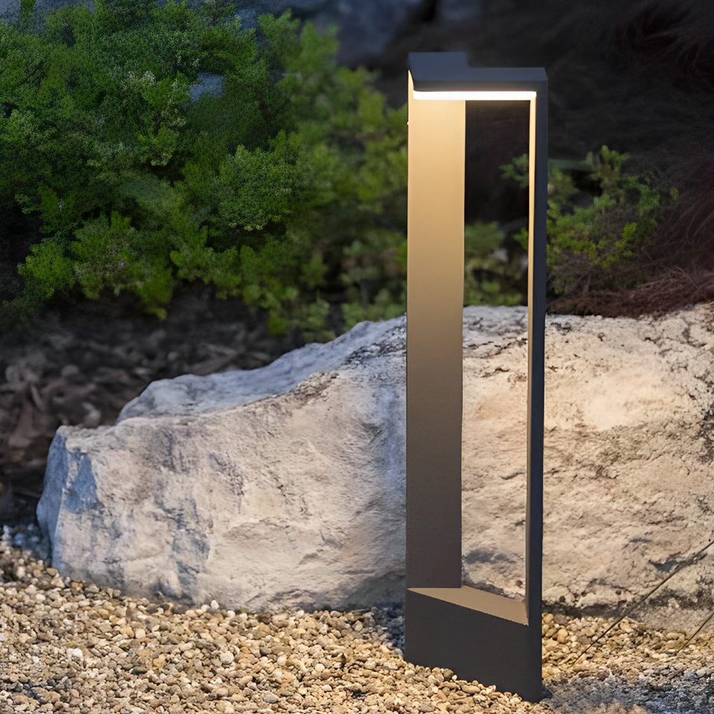 Sleek architectural garden path light glowing softly next to a large grey rock and green bush.