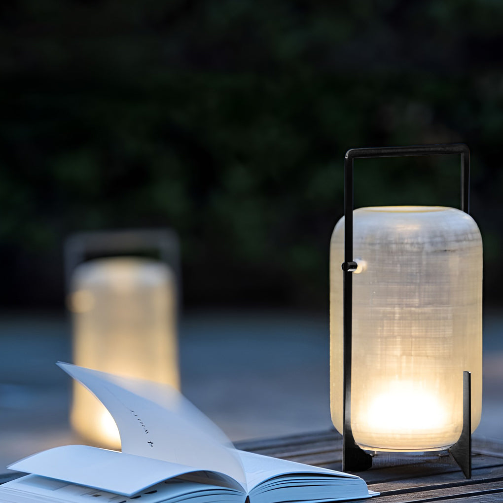 Outdoor modern handle lantern casting soft light on wooden deck with lush green background.