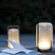 Outdoor modern handle lantern casting soft light on wooden deck with lush green background.