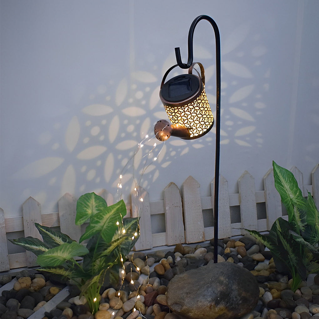 Solar watering can light casting decorative patterns beside plants and a white garden fence.