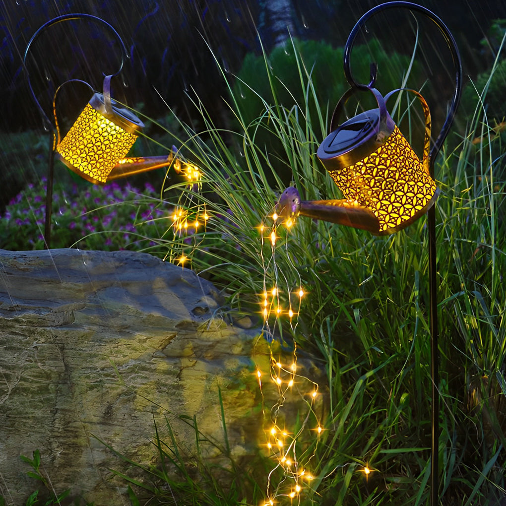 Solar watering can light illuminating ornamental grass and a garden rock with flowing fairy lights.
