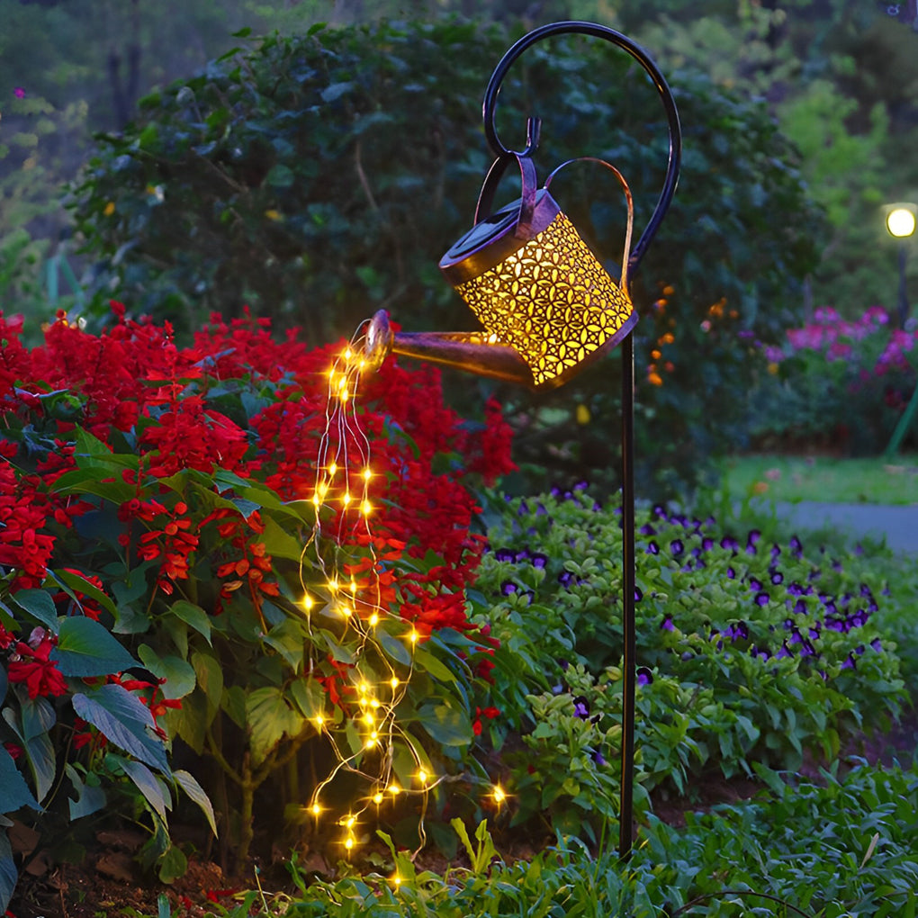 Solar watering can light creating a warm cascading glow above red flowers in an outdoor garden.
