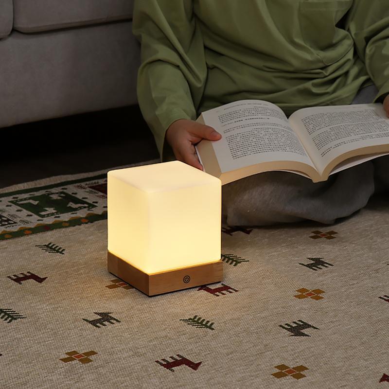 Square wood lamp with a warm glow, placed on a wooden table with a book.