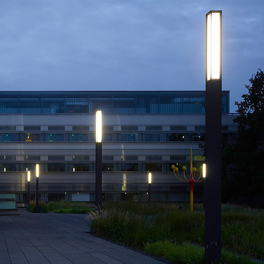 Tall modern black outdoor bollard lights illuminating a paved stone walkway at a large campus.