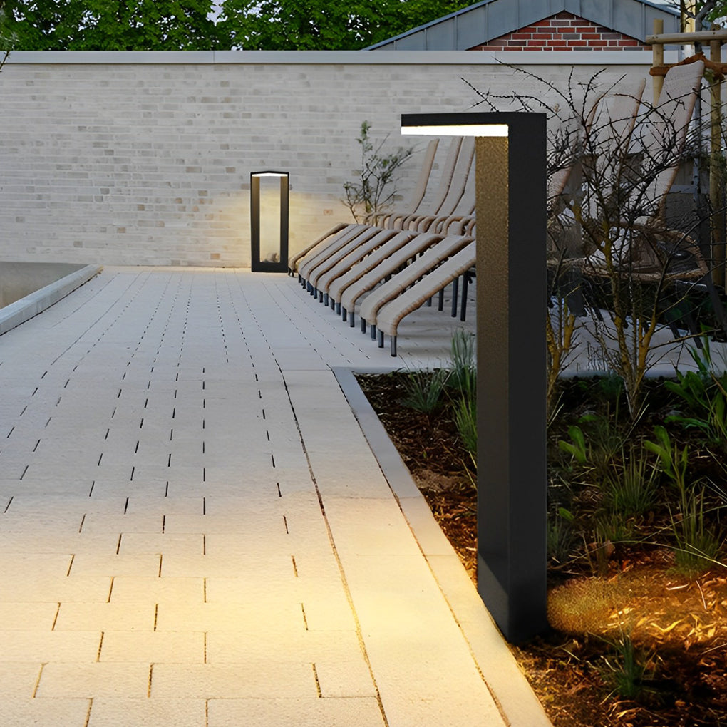 Two modern architectural garden path lights glowing near lounge chairs on a stone patio area ok.